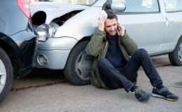 A male car accident survivor sitting by his damaged vehicle, reflecting on the impact of the incident.